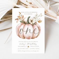 a white pumpkin birthday party card with flowers and greenery on the front, sitting next to some feathers