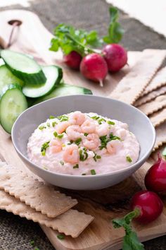 a white bowl filled with food next to crackers and cucumbers on a cutting board