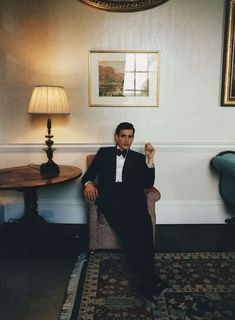 a man in a tuxedo sitting on a chair next to a table and lamp