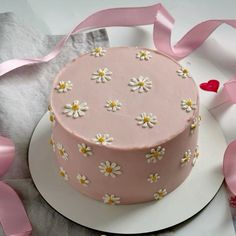 a pink cake decorated with daisies on a white platter surrounded by pink ribbons