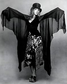 a woman in a black dress and hat with her arms outstretched