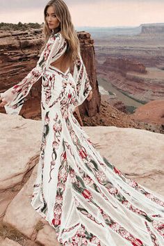 a woman standing on top of a cliff wearing a long white dress with red flowers