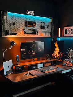 A moody and organized desk setup featuring warm lighting, a central monitor displaying a fire scene, surrounded by tech gadgets and plants. The workspace includes a pegboard organizer with various accessories and modern tech devices. Set Up Pc, Work Wallpaper, Trading Room, Earthy Vibes, Mac Setup, Gaming Desk Setup, Cozy Desk