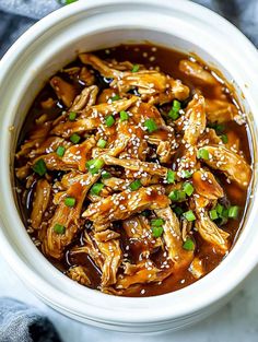 a white bowl filled with chicken and sesame seeds covered in gravy, garnished with green onions