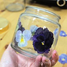 a person holding a glass jar with purple and blue pansies in it on a wooden table