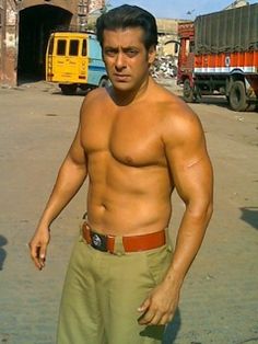 a man without a shirt standing in the middle of an open area with trucks behind him