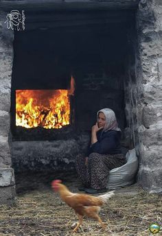 a woman sitting in front of an open fire with a chicken walking past her and talking on the phone