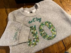 a gray sweatshirt with green and white flowers on it that says art by 10 o'clock