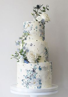 a three tiered white cake with blue and white flowers on the top, sitting on a stand