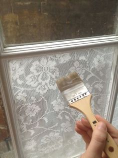 a person holding a paint brush in front of a window with white lace on it