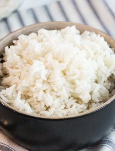 white rice is in a black bowl on a striped table cloth next to a fork