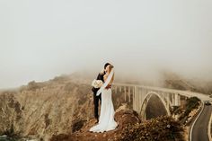 a bride and groom standing on the edge of a cliff with a bridge in the background