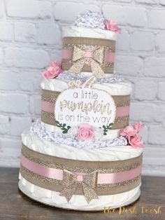 a three tiered cake decorated with pink and brown ribbons, flowers, and a little pumpkin on the way sign