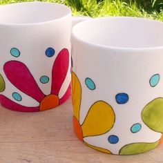 two colorful cups sitting on top of a wooden table next to each other and grass in the background