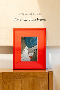 a red frame sitting on top of a wooden dresser next to a wall with a sign that reads, tone - on - tone frame