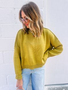 a woman standing in front of a white brick wall wearing a yellow sweater and jeans