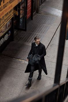 a man is walking down the street with a coat over his shoulders and a bag on his shoulder