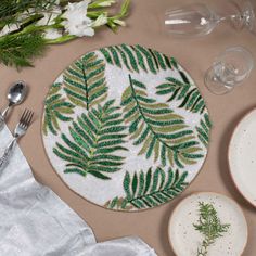 a place mat with green leaves on it next to plates and silverware, wine glasses and flowers