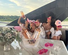 four women sitting at a table with hats and sunglasses on, one holding up a champagne bottle