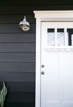 a white door sitting next to a green plant on the side of a building with a light hanging above it