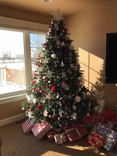 a christmas tree with presents under it in front of a large window and a flat screen tv