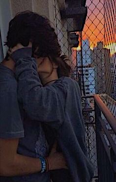 two people embracing each other while standing on a balcony overlooking the cityscape at sunset