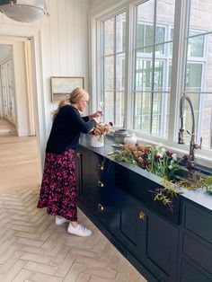 a woman standing in front of a kitchen sink with flowers on the counter and windows