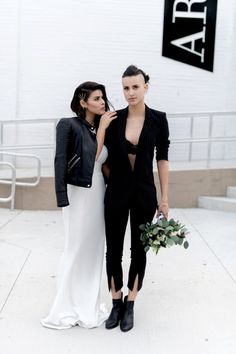 two women standing next to each other in front of a white building with a black sign