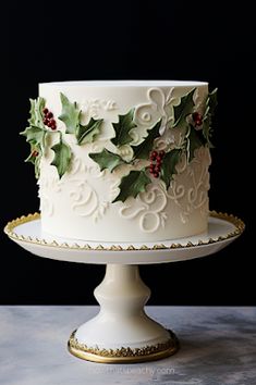 a white cake decorated with holly leaves and red berries sits on a gold platter