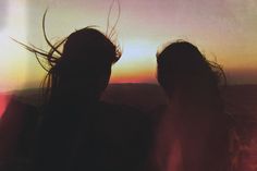 two people standing in front of a sunset with long hair blowing in the wind and one person holding a cell phone up to their ear