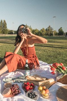 a woman is sitting on the grass with food and drinks in front of her, taking a photo