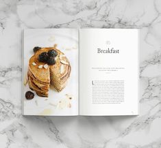 an open cookbook with pancakes and blackberries on the cover is sitting on a marble countertop