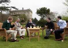 four men are sitting on wicker chairs in the grass, talking and having drinks