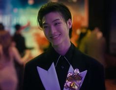 a young man in a black shirt and purple bow tie smiles at the camera while people are dancing behind him