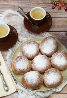 powdered sugar covered donuts on a plate with two cups of tea and spoons