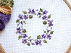 a cross - stitch wreath with purple flowers and green leaves is next to a ball of yarn