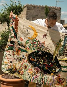 a man holding up a large piece of cloth with pictures on it and plants in the background