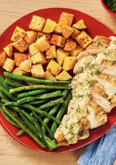 a red plate topped with chicken, potatoes and green beans on top of a wooden table