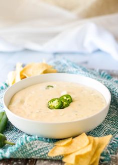 a white bowl filled with dip surrounded by chips and jalapenos on a blue towel