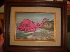 an octopus painting is displayed in a wooden frame on the table next to a person's hand