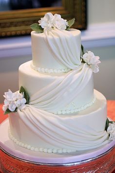 a three tiered white wedding cake with flowers on top is sitting on a glass platter