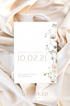 the wedding card is laying on top of white satin with pink flowers and greenery