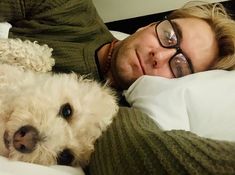 a man laying in bed next to a white dog and wearing eyeglasses with his head on the pillow