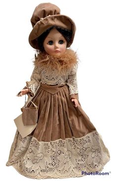 a doll is dressed in a brown dress and hat with lace on the skirt, holding a purse