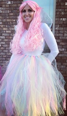 a woman with pink hair wearing a white dress and pastel wig, standing in front of a brick wall