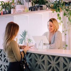 two women sitting at a counter in front of a white wall with plants on it