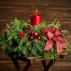 An arrangement made of noble fir, cedar, and white pine with natural pinecones, red berry clusters, red ball ornaments, a glittery red bow, and a metallic red pillar candle with a dark wood background. Christmas Lantern Centerpieces, Fresh Christmas Wreaths, Red Ball Ornaments, Red Candle, Lantern Centerpieces, Light The Way, Candle Ring, Santa's Little Helper