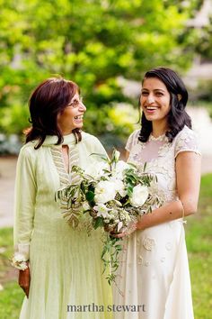 two women standing next to each other in front of trees and grass, one holding a bouquet