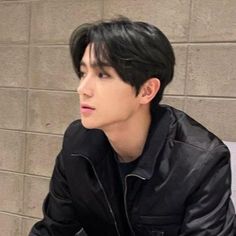 a young man sitting on top of a white bench next to a brick wall wearing a black jacket