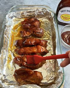 a person holding a brush over some chicken wings in a baking pan with sauce on the side